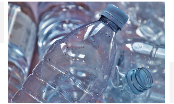 A close-up of several transparent plastic water bottles with blue caps, some lying horizontally and others standing vertically. Droplets of water are visible on the surface of the bottles.