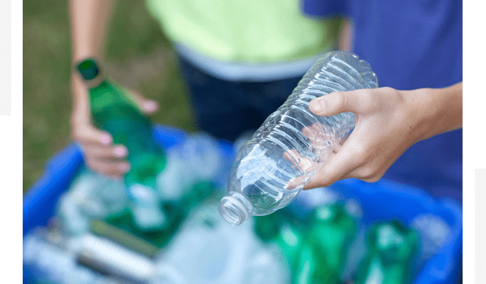 Hands holding plastic and glass bottles over a recycling bin filled with various bottles.